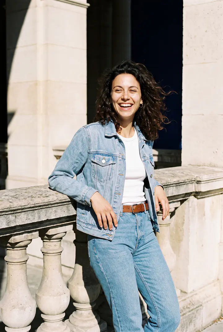 Eine junge Frau in Jeansjacke und Jeans lehnt lachend an einer Steinbalustrade.